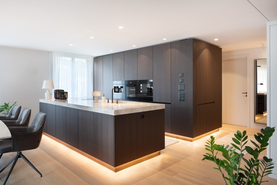 Moderner, offener Küchenbereich mit einer großen Kücheninsel aus dunklem Holz und einer hellen Marmor-Arbeitsplatte. Die Küche ist minimalistisch gestaltet, mit eingebauten schwarzen Geräten und indirekter Beleuchtung unter der Kücheninsel und den Schränken. Im Vordergrund stehen zwei graue Stühle mit Armlehnen. Der Raum ist hell durch große Fenster mit weißen Vorhängen, und es sind einige grüne Pflanzen im Raum verteilt. Der Boden ist aus hellem Holz, und die Atmosphäre wirkt warm und einladend.