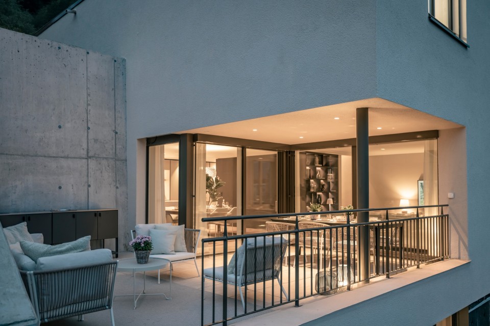 Abendliche Terrasse eines modernen Hauses von Caso Ritter Immobiliendienstleistungen: überdachter Balkon mit eleganten Lounge- und Essmöbeln, weichen Kissen und Pflanzen; durch große Glasfronten Blick in das warm beleuchtete Wohnzimmer mit Regalwand. Schlanke Metallgeländer, dezente Einbaubeleuchtung und klare Architektur neben einer Betonwand schaffen eine ruhige, hochwertige Atmosphäre.