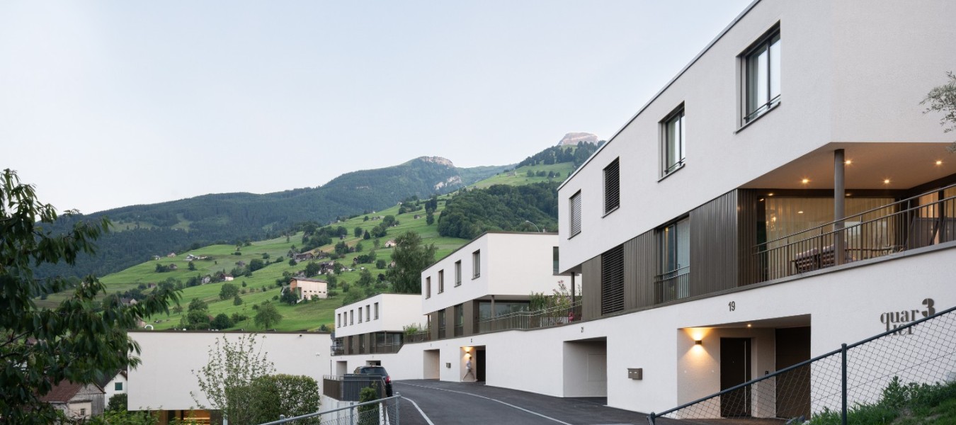 Aussenansicht der modernen Wohnüberbauung ‚Quartier 3‘ in Grabs in der Abenddämmerung. Das Bild zeigt eine Reihe von weissen, minimalistischen Doppeleinfamilienhäusern, die staffelförmig an einem grünen Hang angeordnet sind. Die Gebäude verfügen über Flachdächer, dunkle Fensterrahmen und warm beleuchtete Loggien. Im Vordergrund führt eine kurvige Asphaltstrasse an den Häusern vorbei, während im Hintergrund eine bergige Landschaft unter einem hellen Himmel zu sehen ist.