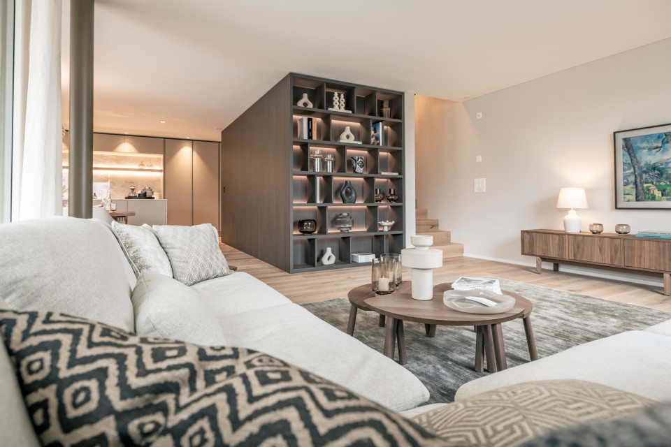 Modernes, helles Wohnzimmer von Caso Ritter Immobiliendienstleistungen: grosse cremefarbene Eckcouch im Vordergrund, zwei runde Holztische mit Vasen und Gläsern auf grauem Teppich, zentrale wandhohe Regalwand mit indirekter Beleuchtung, Keramikobjekten und Büchern; rechts ein niedriges Holz-Sideboard mit Tischlampe und gerahmtem Bild, im Hintergrund eine offen gestaltete, warm beleuchtete Küche sowie eine kleine Treppe und helle Holzfussböden.